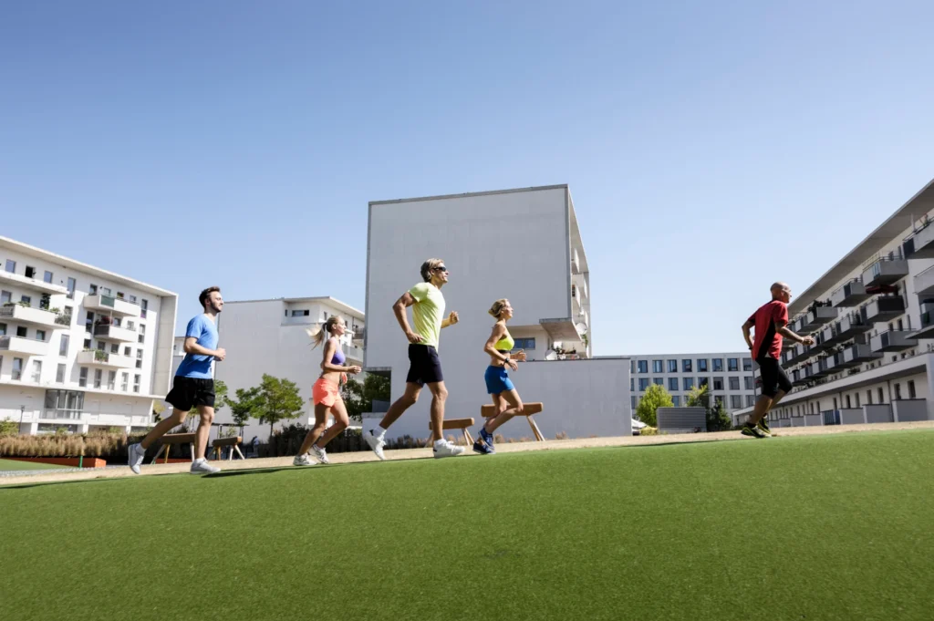 A fitness trainer leading a small group of adults on a run across a synthetic turf surface in an urban courtyard on a sunny day