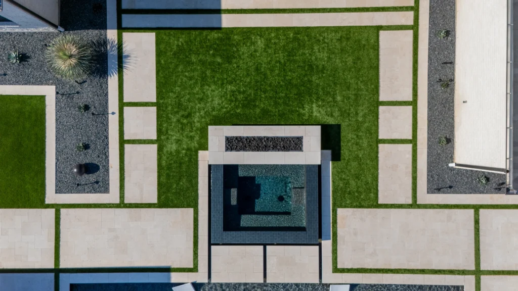 Aerial view of modern backyard with dark-tiled square swimming pool and integrated fire pit, surrounded by vibrant green synthetic turf, light-colored paving stones, and dark gravel beds with desert plants and decorative elements.