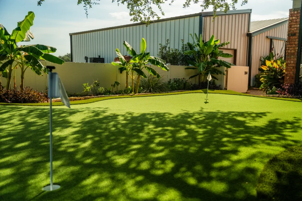 Backyard with vibrant green synthetic turf putting green featuring two white golf flags, surrounded by lush banana trees and other foliage, with corrugated metal buildings and a white privacy wall in the background, under a bright sky with dappled tree shadows.